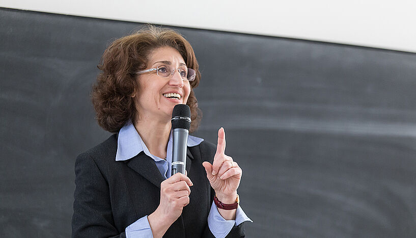 Eine Professorin steht vor einer Tafel, hält ein Mikrofon in einer Hand und erklärt etwas.