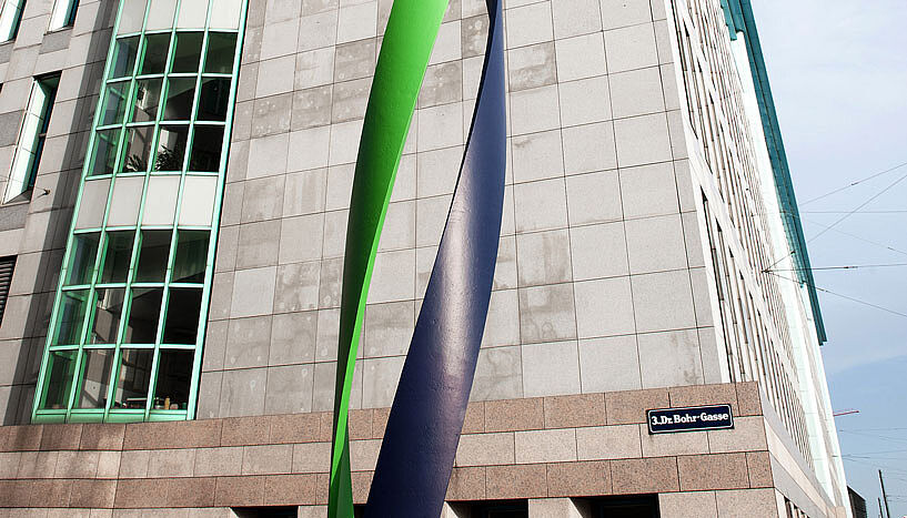 Die Fassade der Max F. Perutz-Laboratories in der Dr. Bohr-Gasse im 3. Bezirk. Es handelt sich um eine beige-braune Fassade, davor befindet sich ein grün-blaues, geschwungenes Kunstwerk, das an einen DNA-Strang erinnert. 