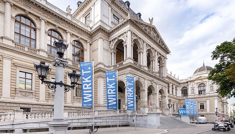 Außenansicht des Hauptgebäudes der Universität Wien mit blauen Flaggen, auf denen 